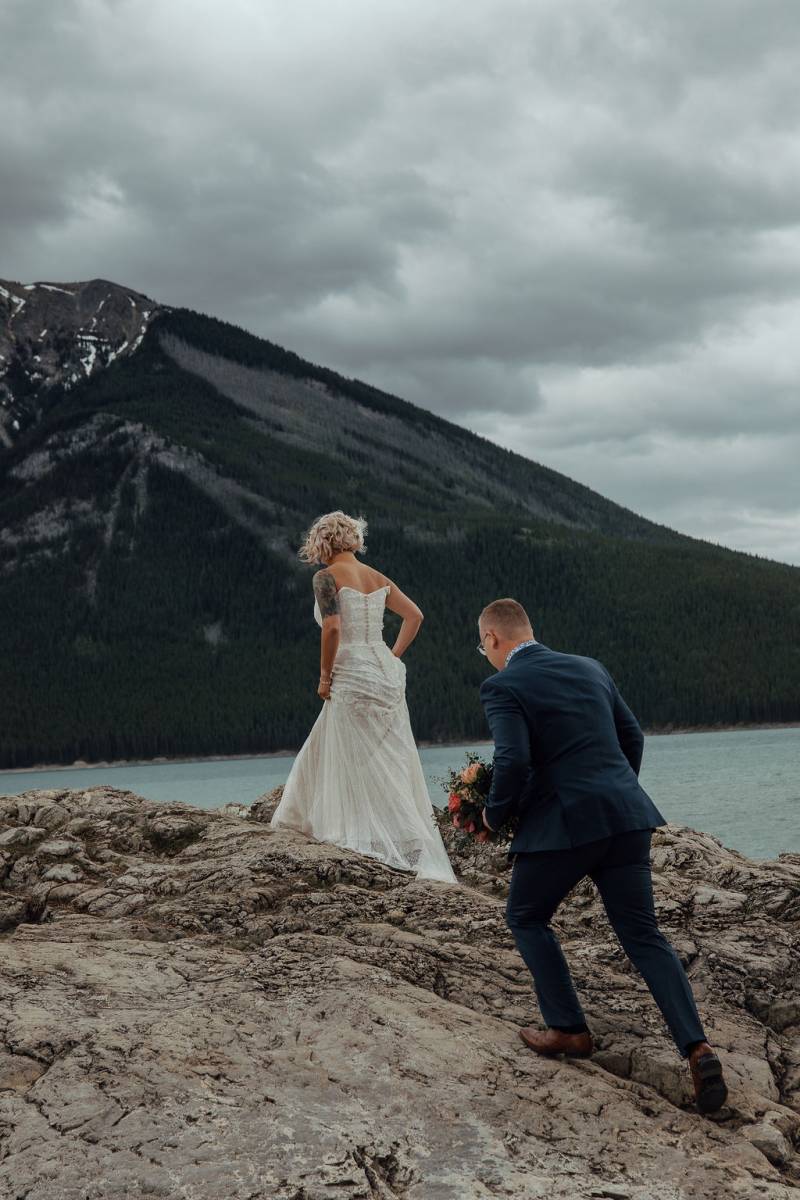 Bride and groom walk up rocky surface overlooking lake and mountains