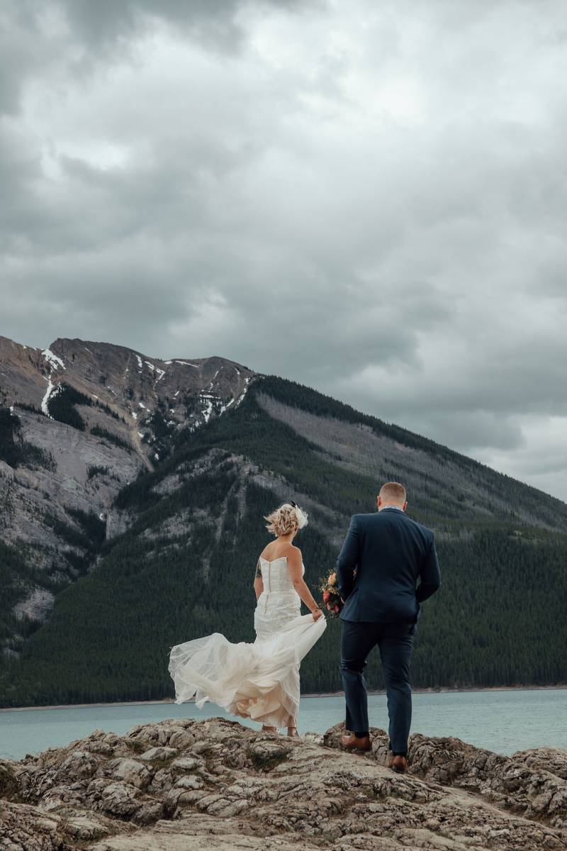 Bride and groom stand on rocky surface wind blowing overlooking lake and mountains 