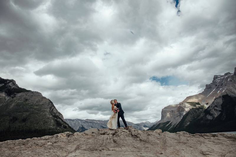 Bride and groom embrace on rocky hill backing mountain peaks 