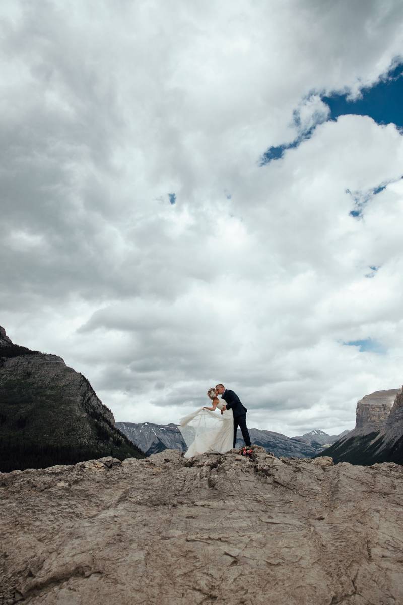 Bride and groom embrace and kiss on rocky hill backing mountain peaks 