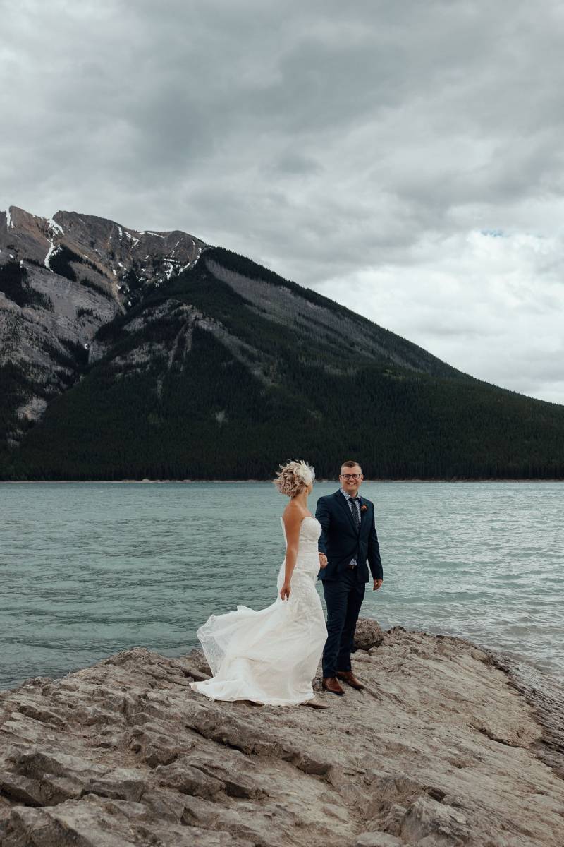 Bride and groom stand smiling on rocky surface overlooking lake and mountains 