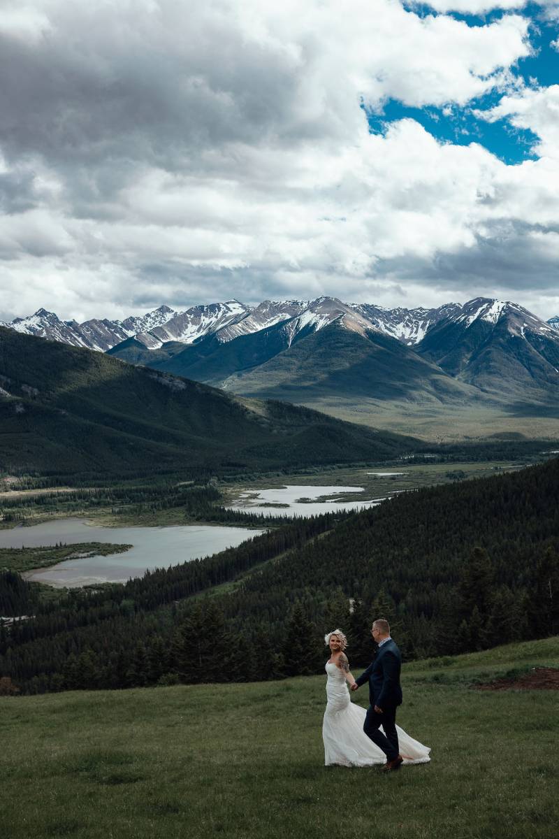 Bride and groom walk holding hands over rolling grassy hills 