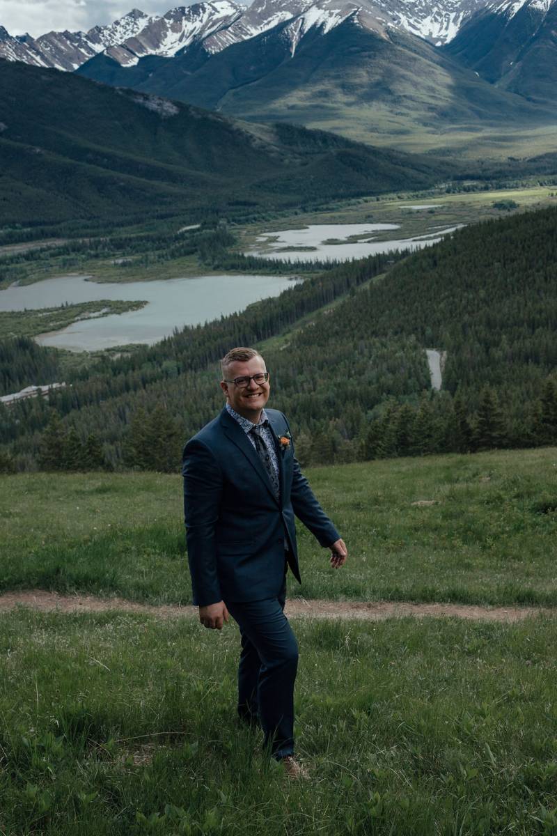 Groom smiling walking up grassy hill overlooking mountains and lake