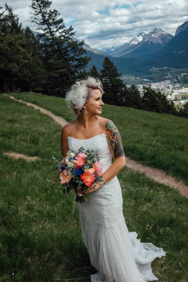 Bride standing on grassy hill holding orange pink and blue bouquet 
