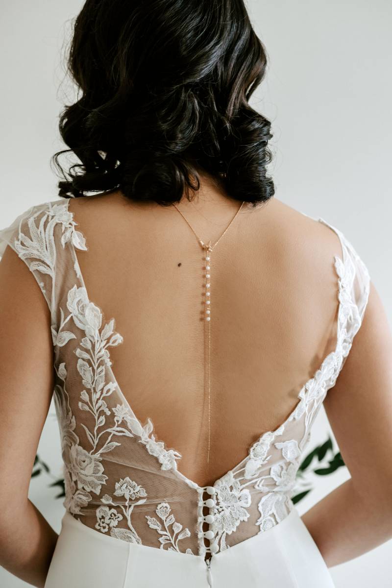 Woman stands facing away in white lace open back dress with necklace hanging 
