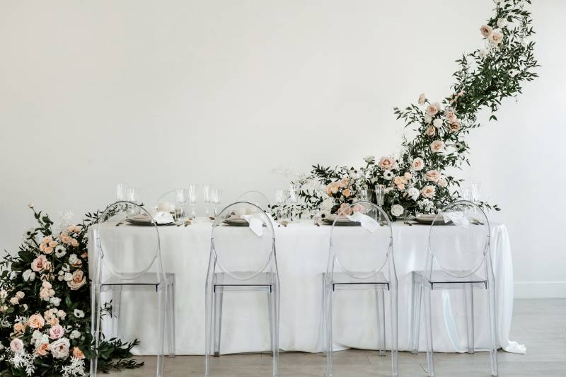 Clear seats around white table and place settings with large blush and white floral arrangement to side 