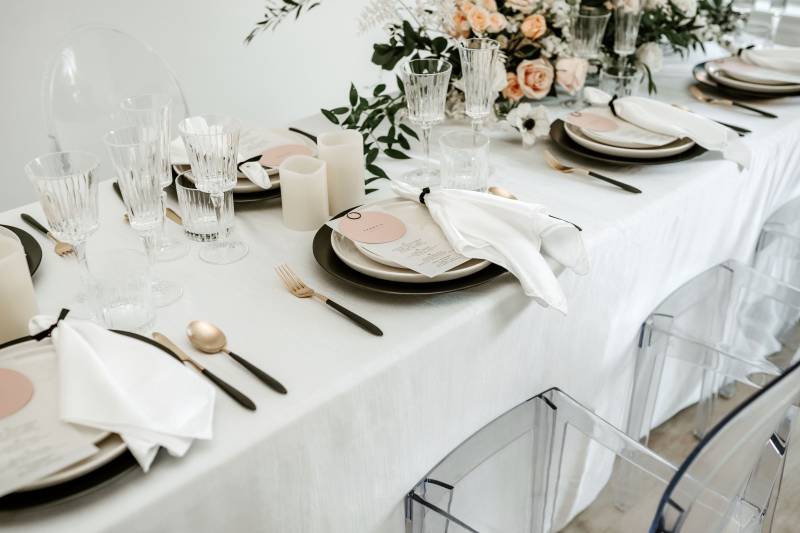 Clear seats around white table and place settings with large blush and white floral centerpiece 