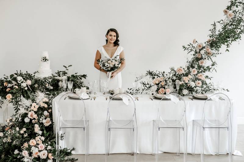 Woman in white dress stands behind white table with clear seats large blush and white floral arrangement centerpiece and to side 