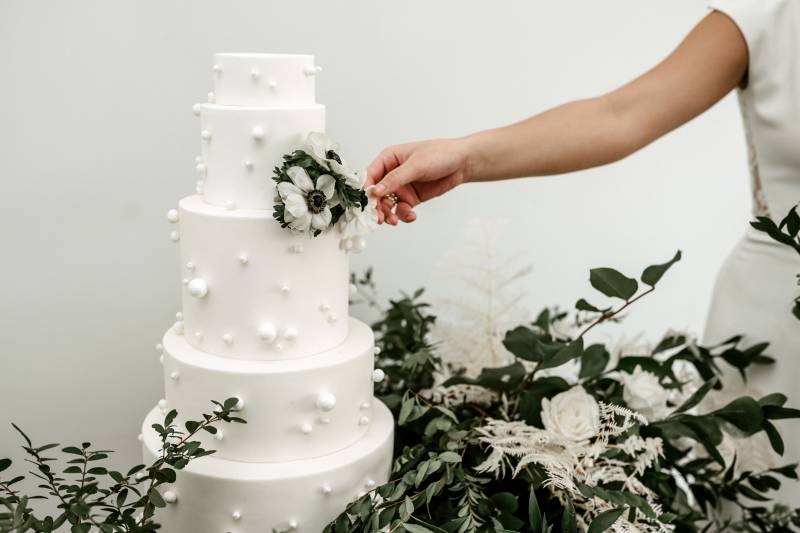 Woman in white dress touches white floral accent on large white wedding cake with greenery surrounding 