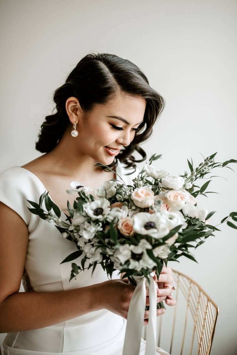 Woman in white dress smiling looking down holding white and blush bouquet and white ribbon