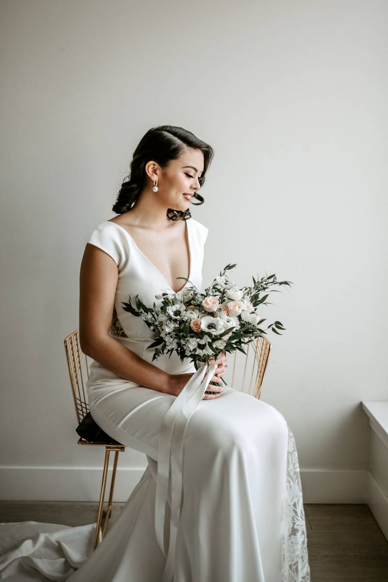 Woman sits in white lace dress holding blush and white bouquet and white ribbon 