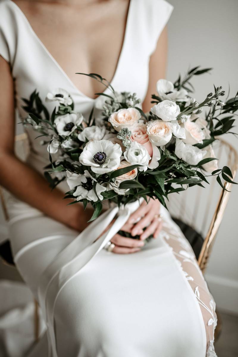 Woman sits in white lace dress holding blush and white bouquet and white ribbon 