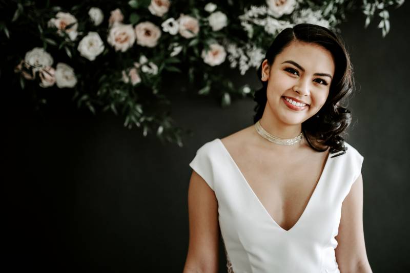 Woman in white dress smiling against black wall with blush and white floral arrangement 