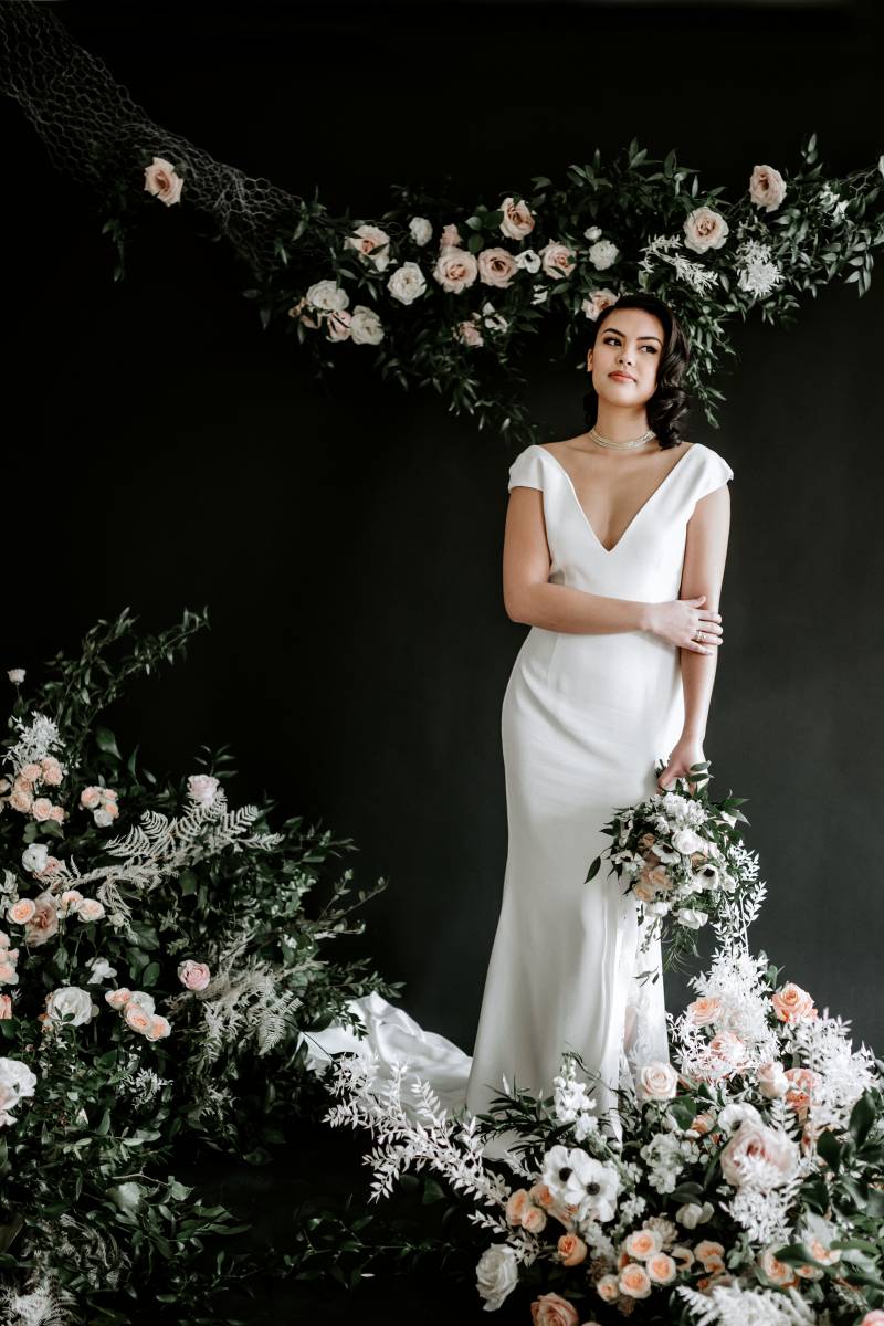 Woman in white dress holds white and blush bouquet to side arm across waist surrounded by blush and white arrangements