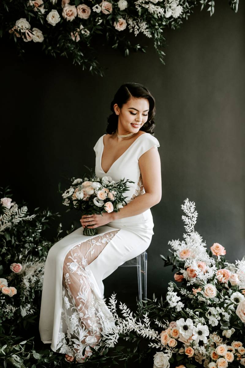 Woman sits surrounded by blush and white floral arrangements in white lace dress holding blush and white bouquet and white ribbon 