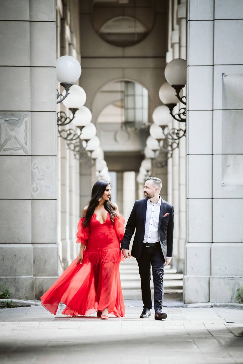 Woman in red dress and man in black suit walk between concrete walls smiling 