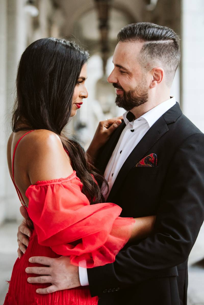 Bride in red shoulder less dress smiling embracing groom in black suit 