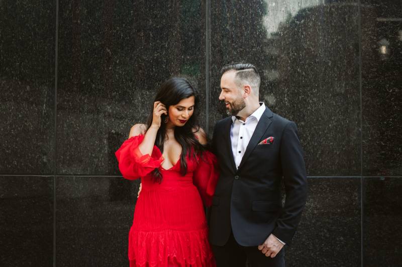 Bride in red shoulder less dress pushes hair behind ear while groom smiles on black wall 