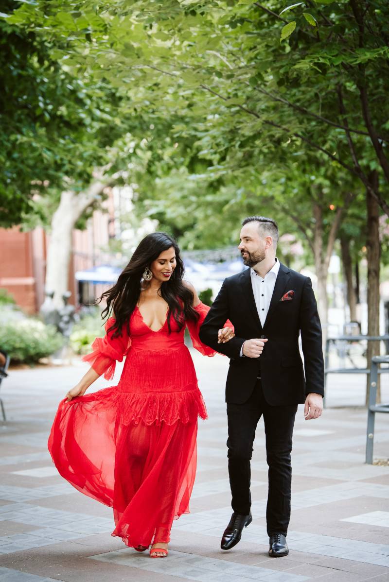 Bride in red dress walks holding arm of groom in black suit down sidewalk 