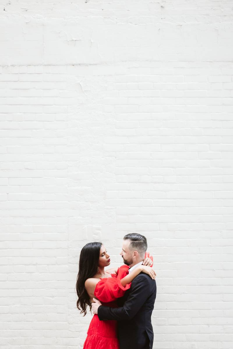 Bride arms over groom shoulder embracing on white brick wall 
