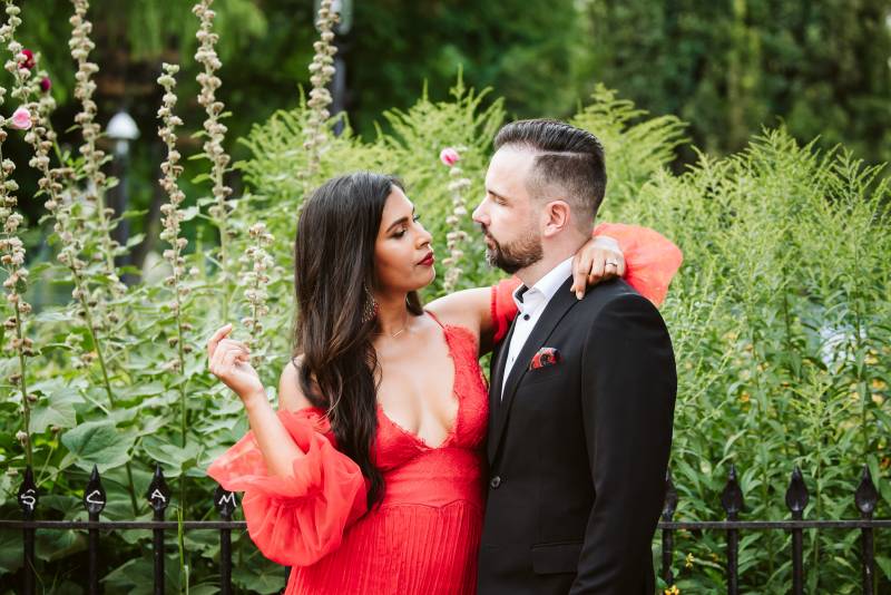 Bride in red shoulder less dress arm around grooms neck in front of greenery 