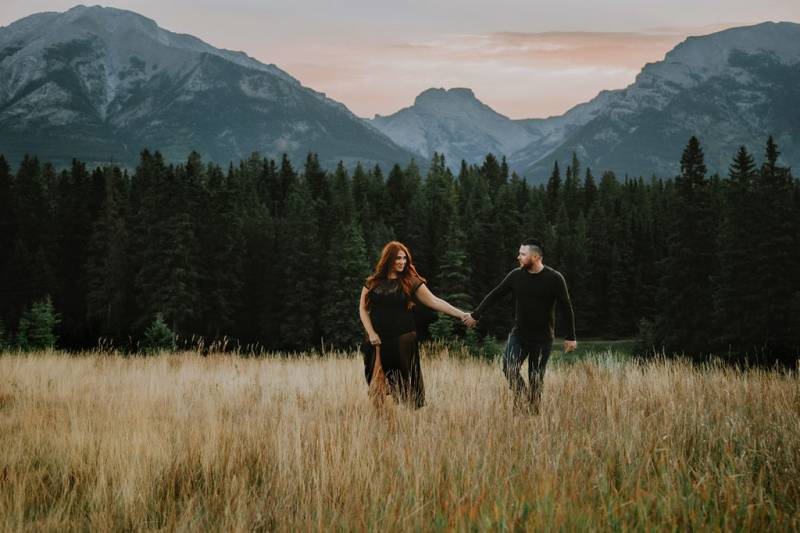 Woman walks in flowing black dress holding hands with man in front of mountains 