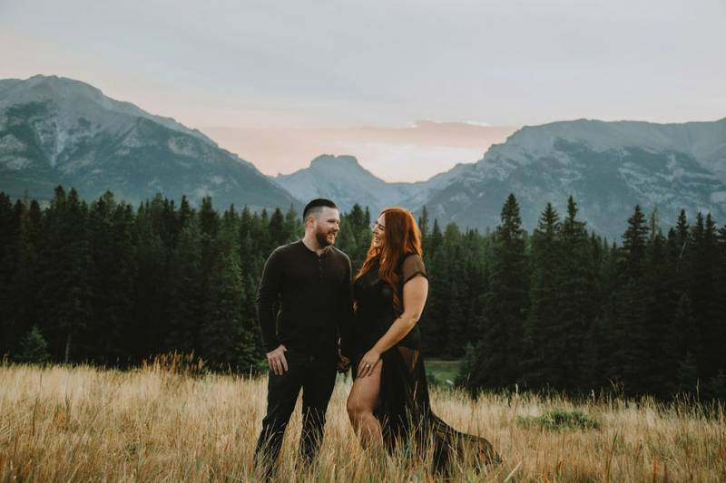 Man and woman stand in brown grassy field laughing in front of mountains 