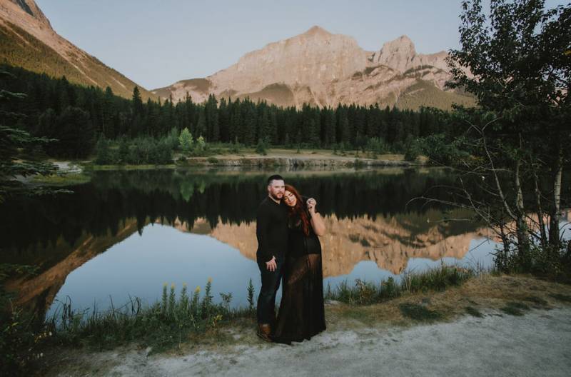 Woman holds mans hand over shoulder in front of river reflecting mountains 