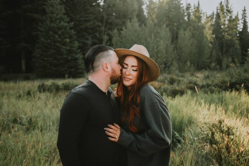 Man kisses woman's cheek smiling in grassy forest field 