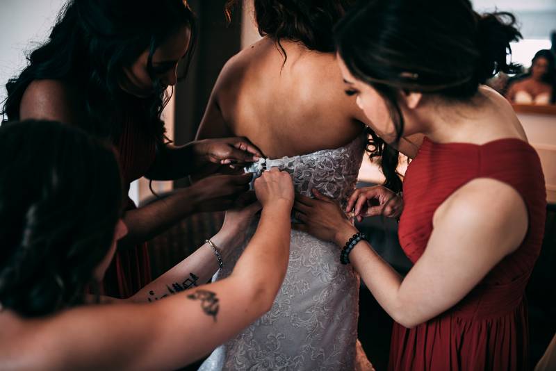 Three women help pin up brides white lace open back dress from behind 