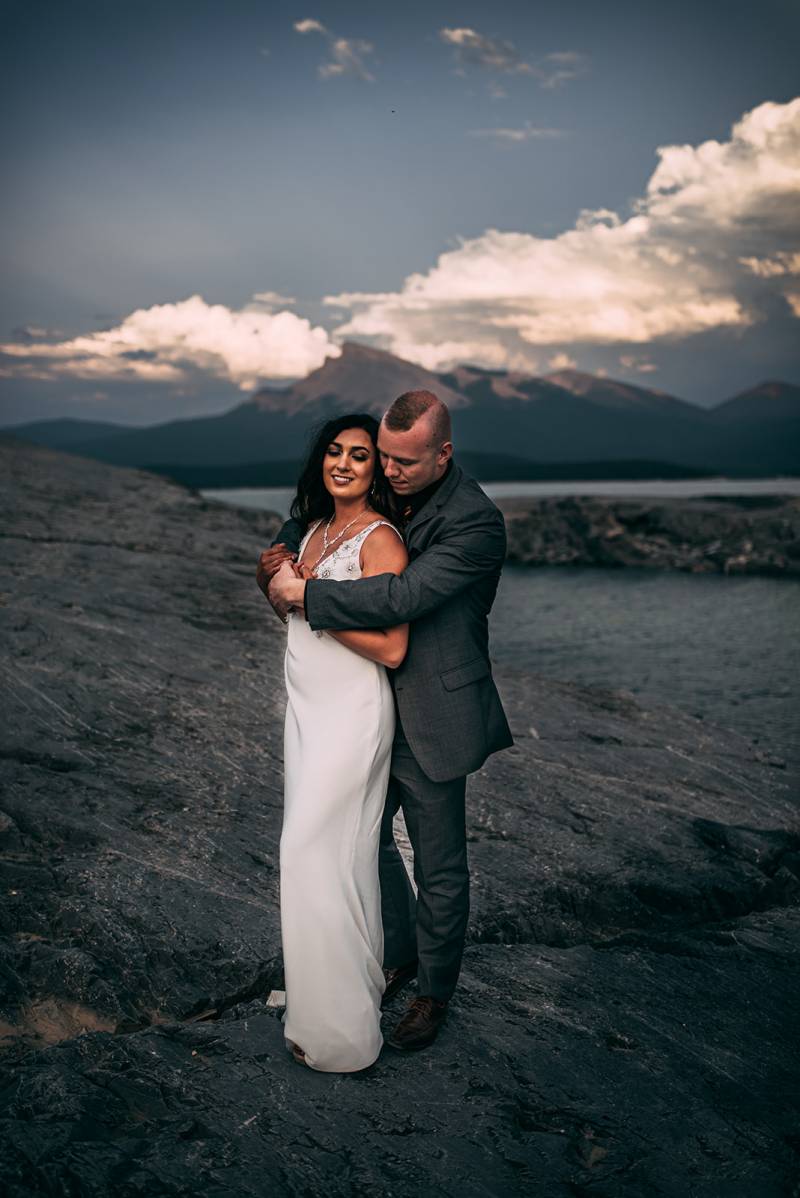 Groom embraces bride from behind standing on rocky slope backing mountains 