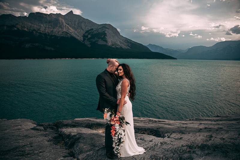 Bride stands embraced by groom holding peach and white bouquet overlooking lake and mountains 