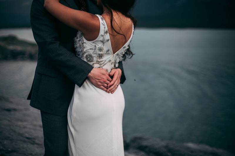 Bride embraced by groom around waist in front of lake  