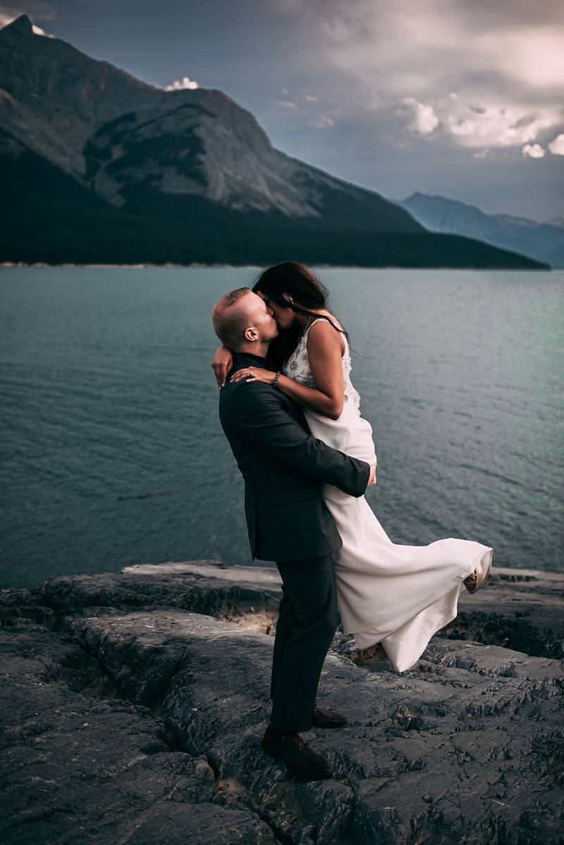 Groom lifts bride kissing on rocky ledge overlooking lake and mountains 