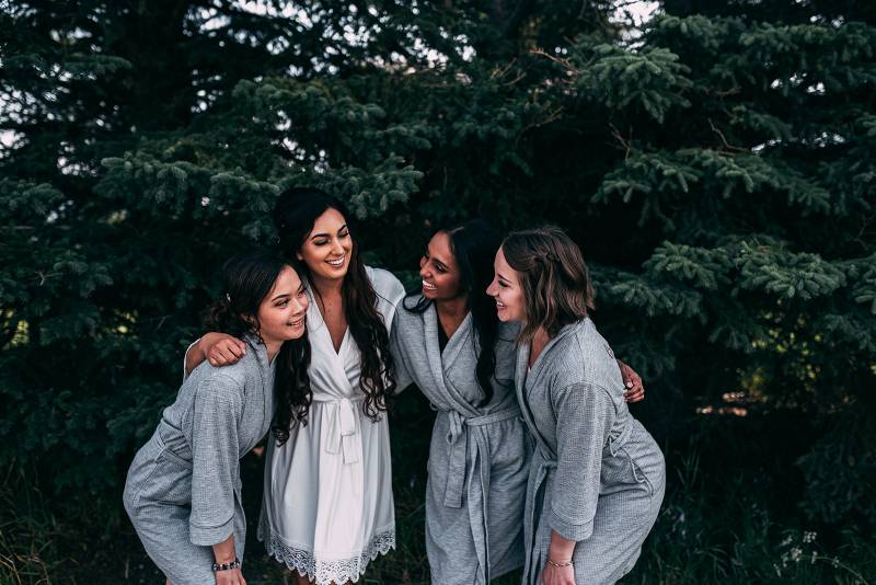 Bride and bridesmaids lean forward smiling arms over shoulders 