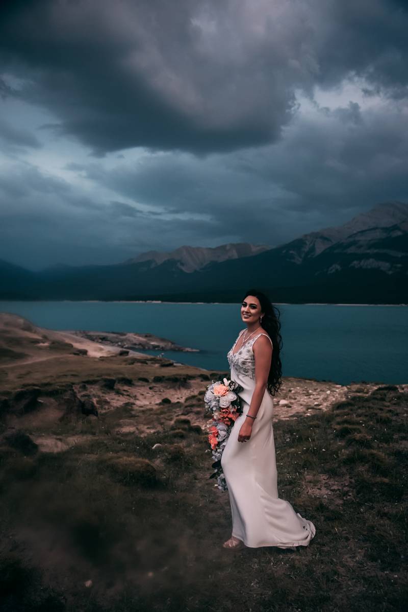 Bride stands on grassy hill in white dress holding peach and white bouquet overlooking lake and mountains 
