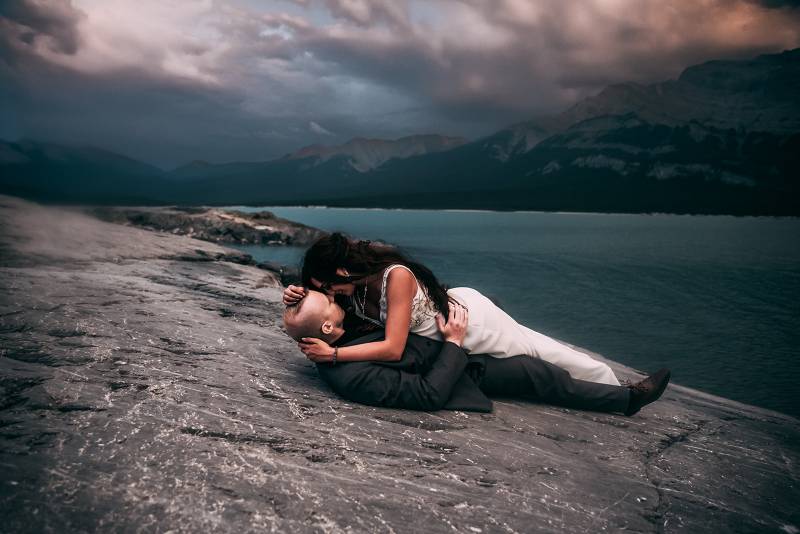 Bride lays on top of groom on rocky slope staring intently overlooking lake and mountains 