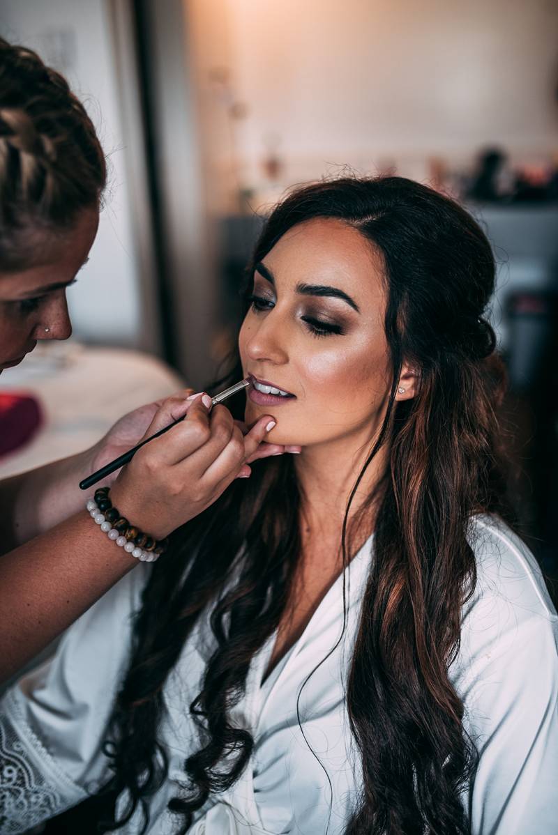 Woman applies lip liner to bride sitting in white robe 