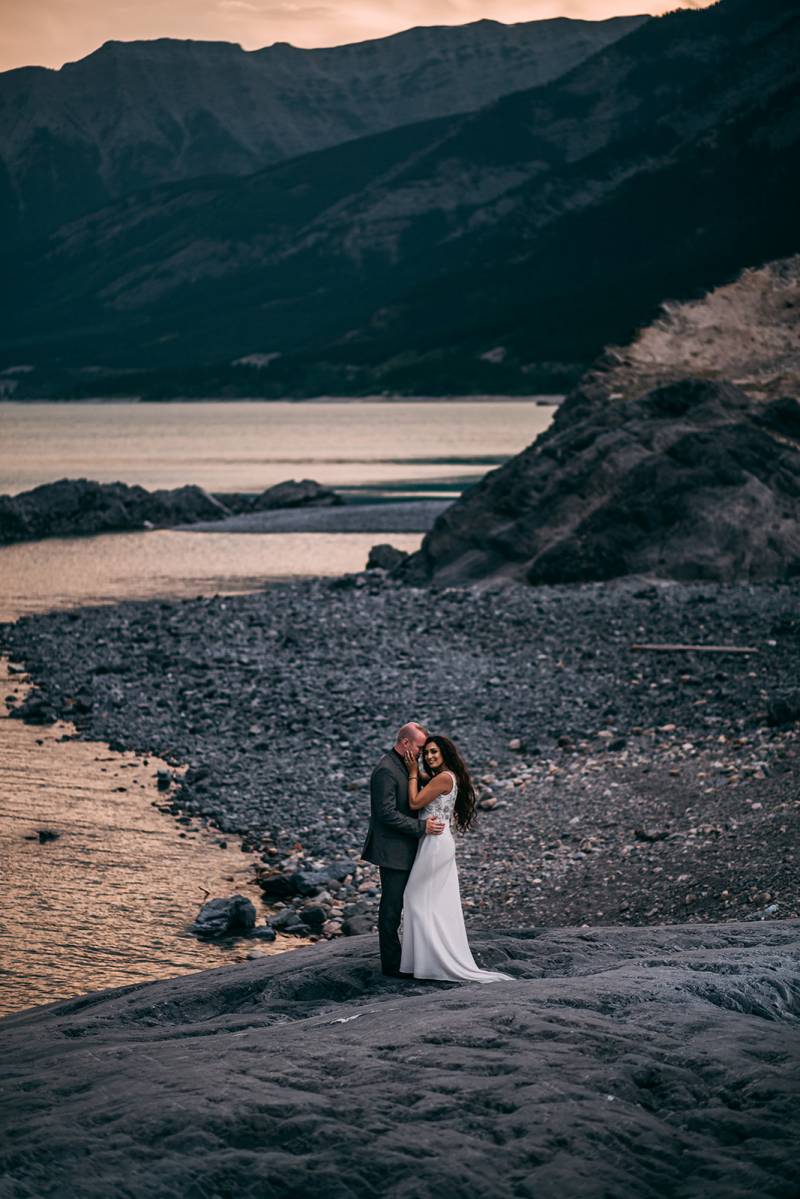 Bride and groom embrace touching foreheads at base of rocky slope overlooking lake and mountains 