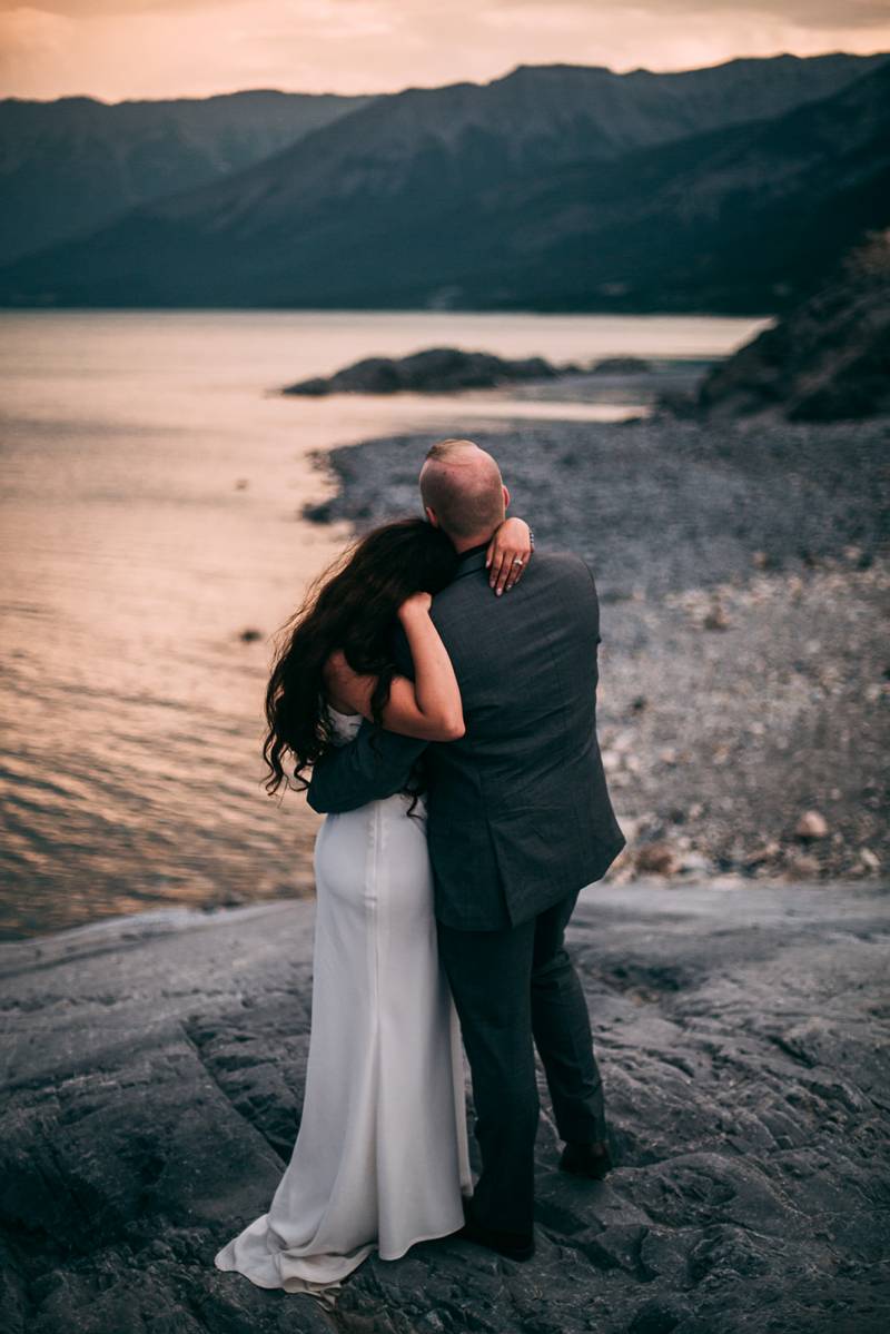 Bride and groom embrace leaning on shoulder on rocky ledge looking at lake and mountains 