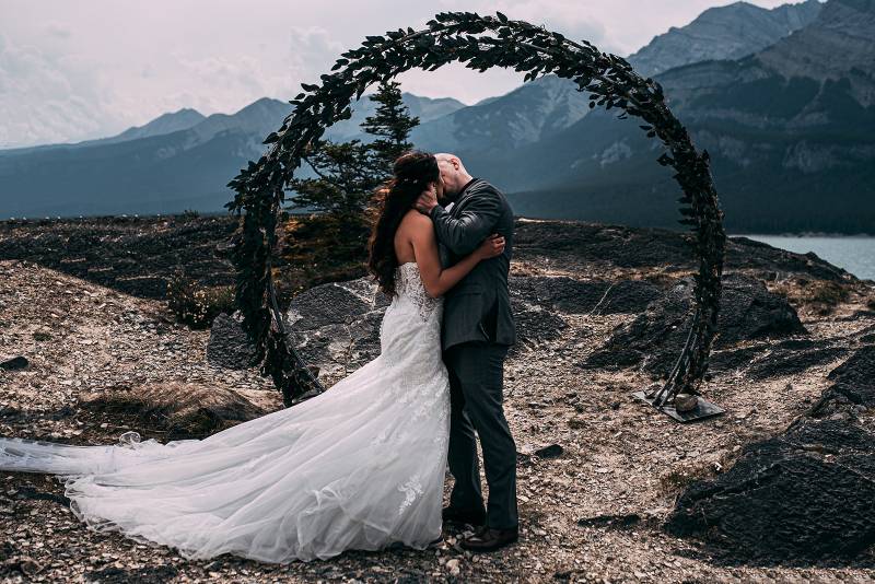 Bride and groom embrace kissing in front of circle pine wedding arch backing mountains 