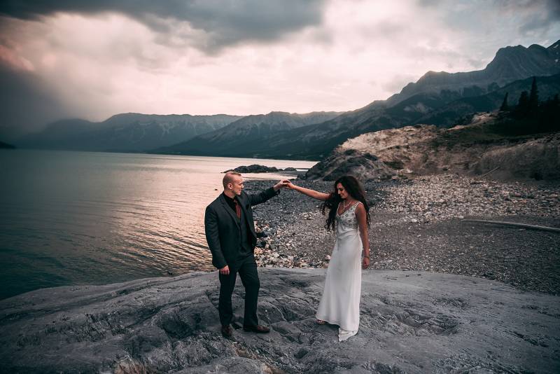 Bride and groom stand holding outstretched arms on rocky slope overlooking lake and mountains 