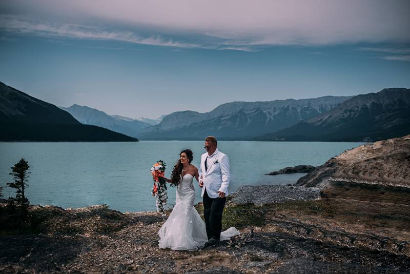 Bride walks in white dress holding peach and white bouquet with father in white suit overlooking lake and mountains 