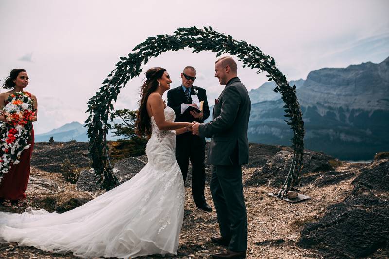 Bride and groom holds hands smiling in front of circle pine wedding arch officiant behind 