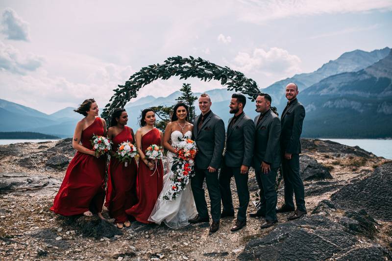 Bride and groom stand in between bridesmaids and groomsmen backing wedding arch and mountains 