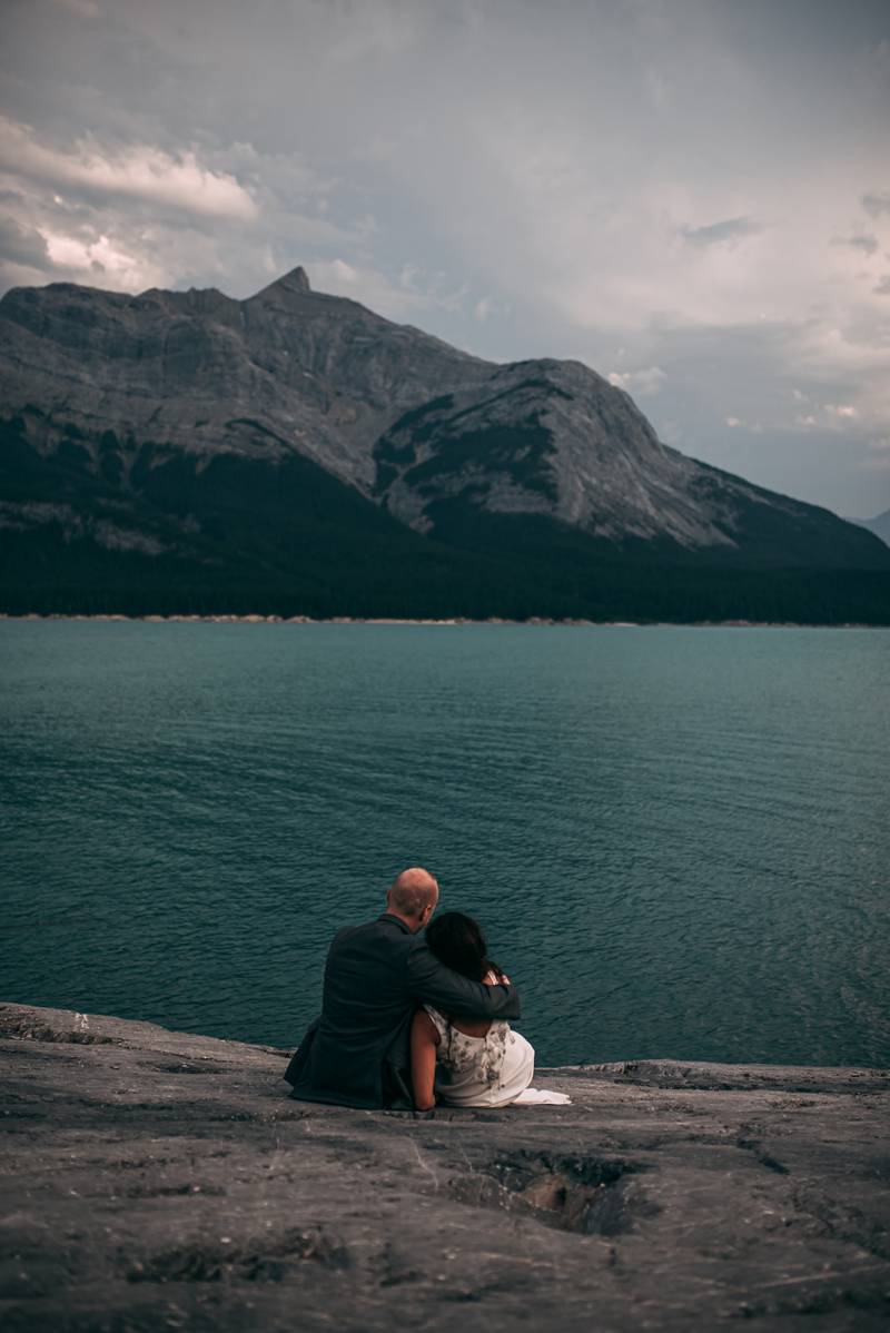 Bride and groom sit arm over shoulder on rocky slope looking onto lake and mountains 