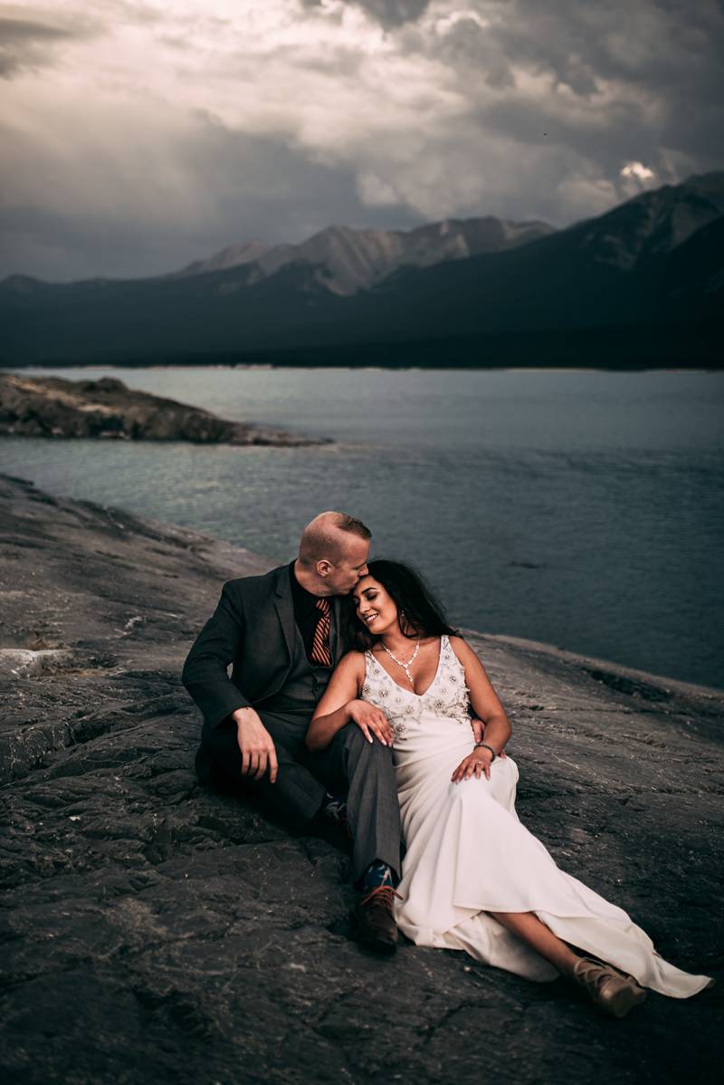 Bride sits leaning on groom kissing forehead on rocky slope overlooking lake and mountains 