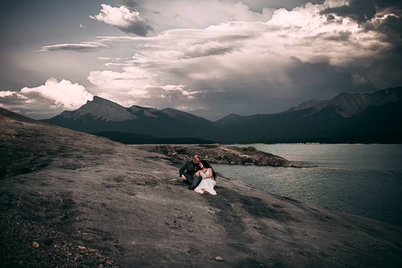Bride sits leaning on groom on rocky slope overlooking lake and mountains 