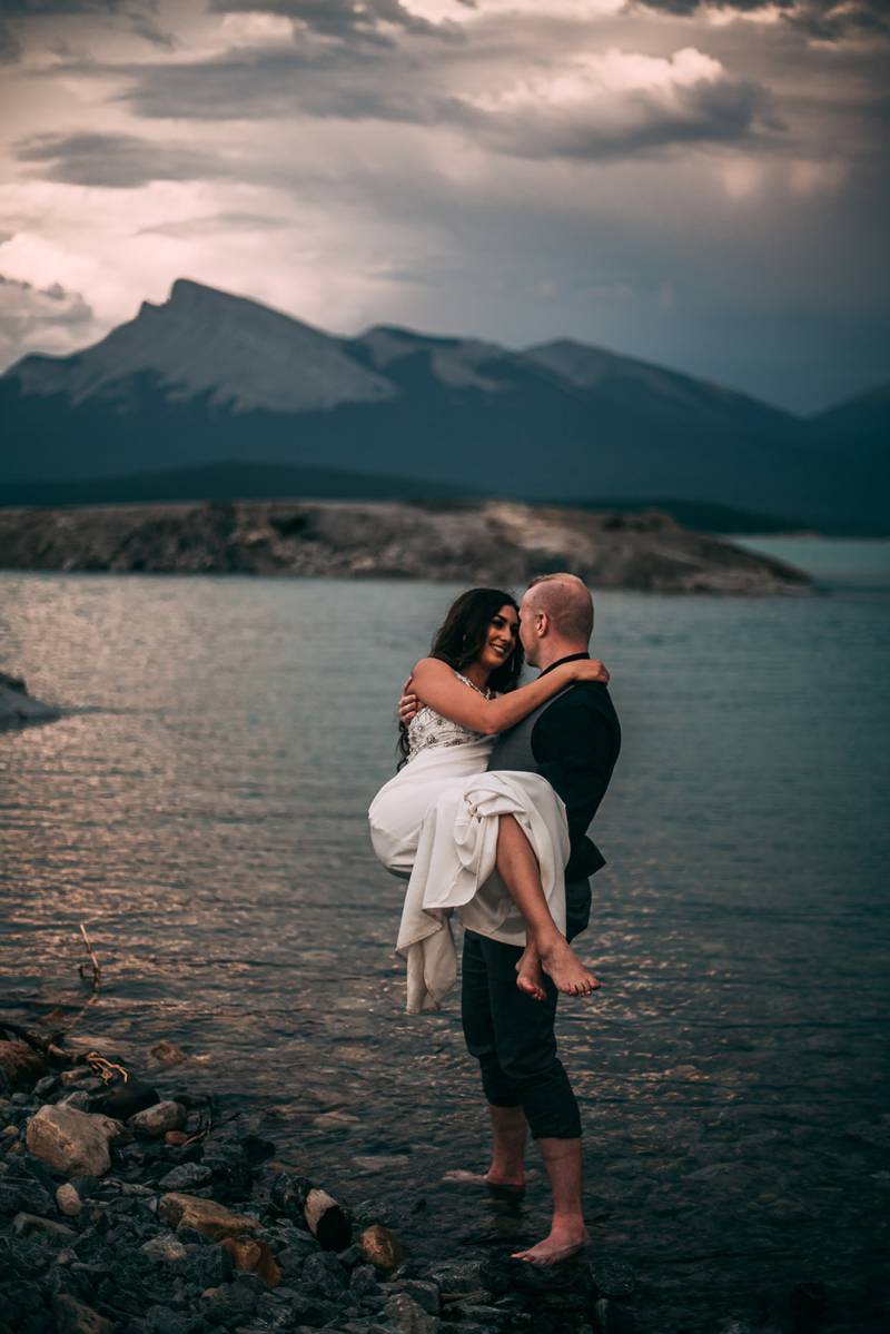 Groom carries bride in pants rolled up in bare feet in lake overlooking mountains 