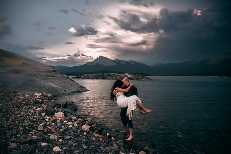 Groom carries bride in pants rolled up in bare feet in lake overlooking mountains 